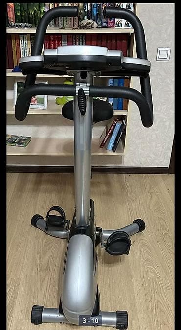 Gümüşü rəngdə, şaquli vəziyyətdə duran stasionar velosiped. Dəstəkləri